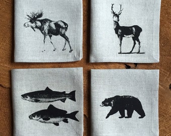 May include: Set of four linen napkins with black and white animal illustrations. The napkins feature a moose, a deer, a salmon, and a bear.