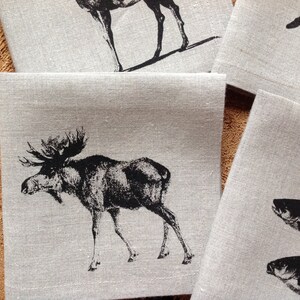 May include: Set of four linen napkins with black and white animal illustrations. The napkins feature a moose, a deer, and two fish.