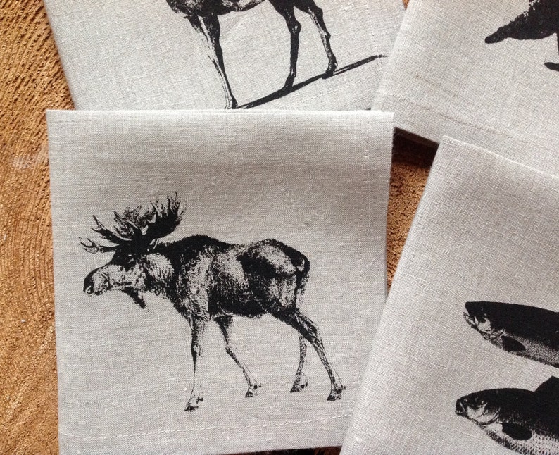 May include: Set of four linen napkins with black and white animal illustrations. The napkins feature a moose, a deer, and two fish.
