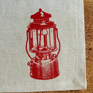 May include: Red vintage lantern printed on a white linen tea towel. The lantern has a round glass globe and a metal handle.