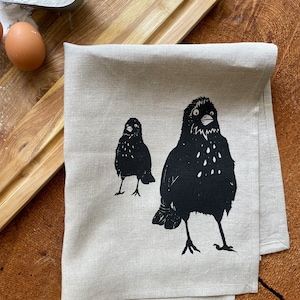 May include: A beige linen kitchen towel with a black and white print of two birds. The birds are facing forward with their beaks open.