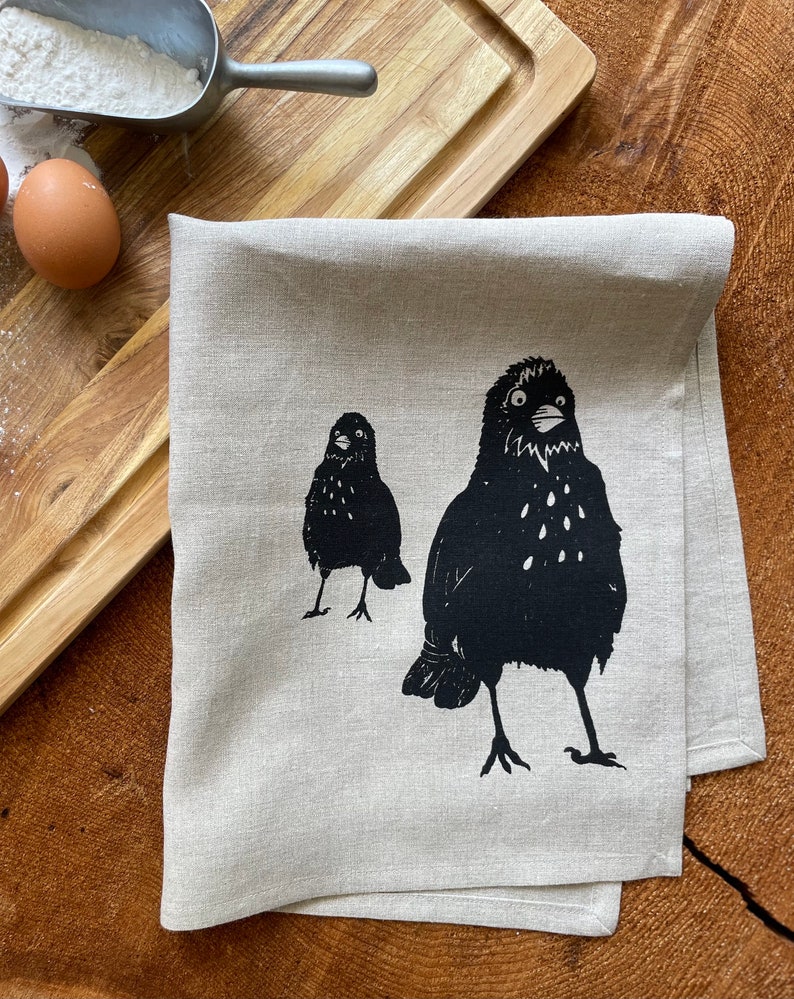 May include: A beige linen kitchen towel with a black and white print of two birds. The birds are facing forward with their beaks open.