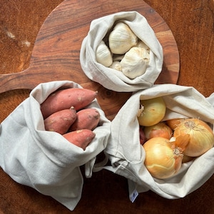 May include: Four reusable produce bags filled with fresh produce. The bags are made of natural linen fabric and are a sustainable alternative to plastic bags. The bags contain garlic, onions, and sweet potatoes.