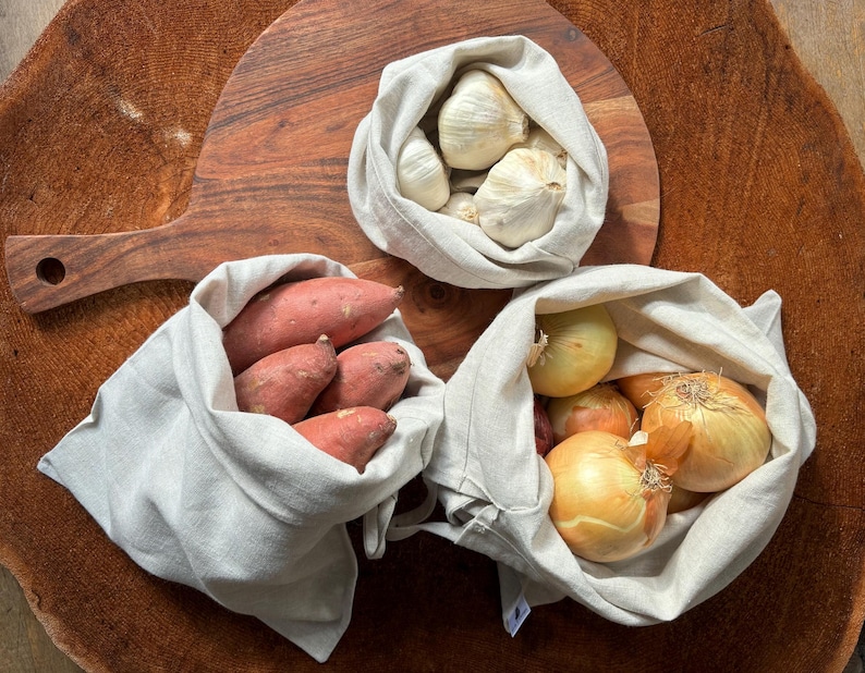 Pode incluir: Quatro sacos reutiliz&aacute;veis para produtos frescos cheios de produtos frescos. Os sacos s&atilde;o feitos de tecido de linho natural e s&atilde;o uma alternativa sustent&aacute;vel aos sacos de pl&aacute;stico. Os sacos cont&ecirc;m alho, cebolas e batatas doces.