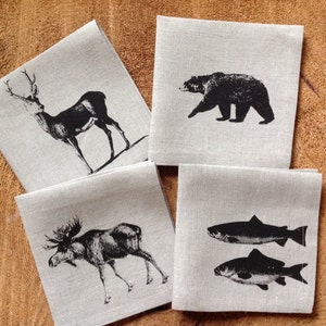 May include: Four linen napkins with black and white illustrations of a deer, a bear, a moose, and two fish. The napkins are on a wooden surface.