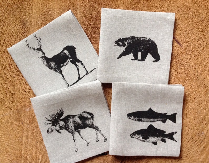 May include: Four linen napkins with black and white illustrations of a deer, a bear, a moose, and two fish. The napkins are on a wooden surface.