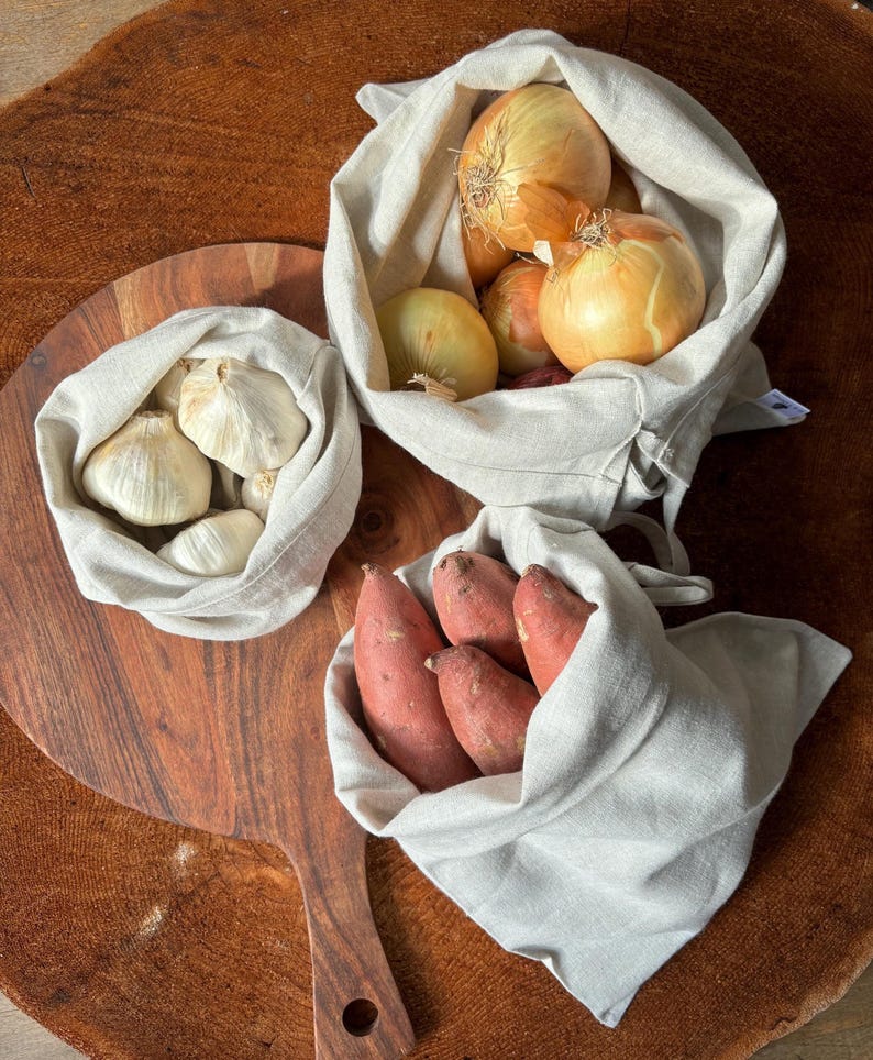Pode incluir: Tr&ecirc;s sacos de tecido reutiliz&aacute;veis ​​cheios de produtos frescos. O primeiro saco cont&eacute;m alho, o segundo cebolas e o terceiro batatas doces.