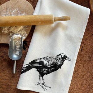 May include: A white linen kitchen towel with a black crow illustration printed on it. The towel is lying on a wooden surface with a rolling pin and a scoop of flour.