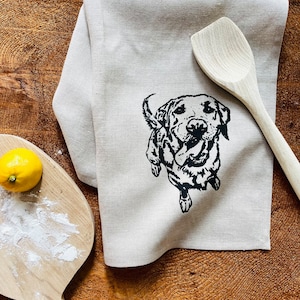 May include: A beige linen kitchen towel with a black and white illustration of a dog printed on it. The towel is folded and lying on a wooden surface.