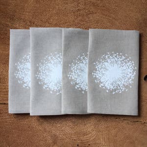 May include: Four folded beige linen napkins with a white dandelion design. The dandelion is centered on each napkin, with a detailed seed head. The napkins are arranged on a wooden surface.