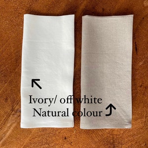 May include: Two linen napkins, one ivory/off white and one natural color. The napkins are folded and placed side-by-side on a wooden surface.