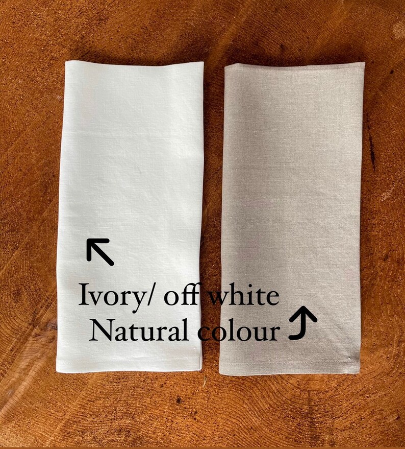 May include: Two linen napkins, one ivory/off white and one natural color. The napkins are folded and placed side-by-side on a wooden surface.