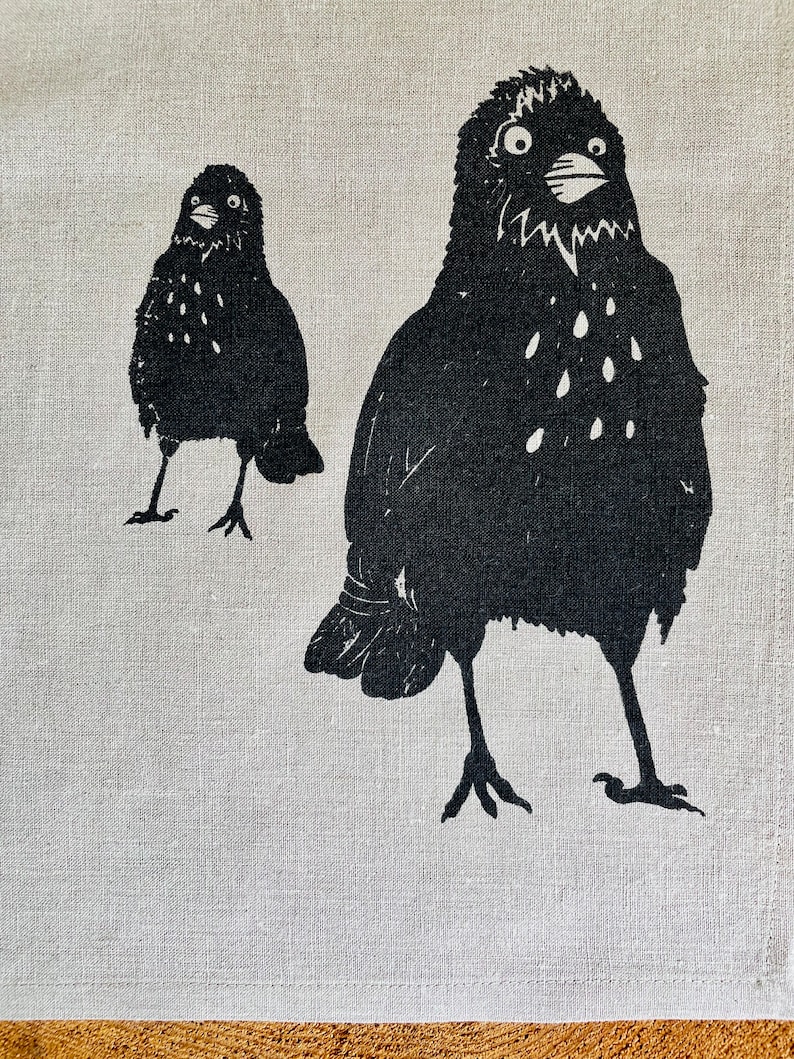 May include: Two black birds with white spots are depicted on a white linen background. The larger bird is facing forward with its beak open, while the smaller bird is facing to the left.