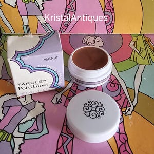 May include: Vintage Yardley Pot o' Gloss lip gloss in the shade Walnut. The open container reveals the brown lip gloss, next to its white lid with a black design. The box is white with a purple and blue design. The net weight is .12 oz.