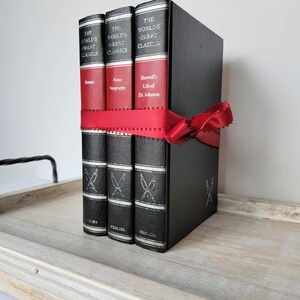 May include: Three black hardcover books with gold lettering and a red ribbon tied around them. The books are titled "The World's Great Classics" and the titles of the individual books are "Essays", "Auto-biography", and "Boswell's Life of Dr. Johnson".