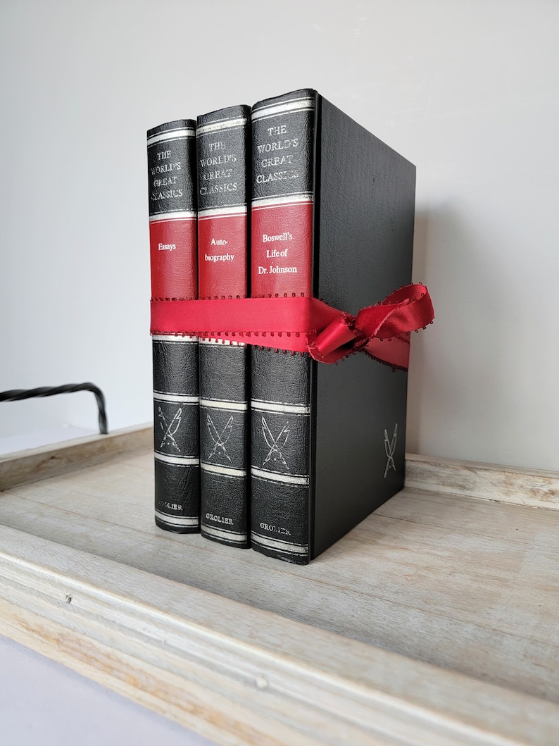 May include: Three black hardcover books with gold lettering and a red ribbon tied around them. The books are titled "The World's Great Classics" and the titles of the individual books are "Essays", "Auto-biography", and "Boswell's Life of Dr. Johnson".