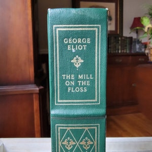 May include: A green hardcover book titled "The Mill on the Floss" by George Eliot. The book's spine features gold lettering and decorative gold accents within a rectangular frame. The book is displayed vertically.