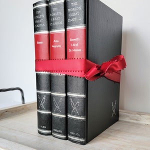 May include: Three black leather-bound books with red spines and gold lettering. The books are titled "The World's Great Classics" and feature the titles "Essays", "Autobiography", and "Boswell's Life of Dr. Johnson". The books are tied together with a red ribbon.