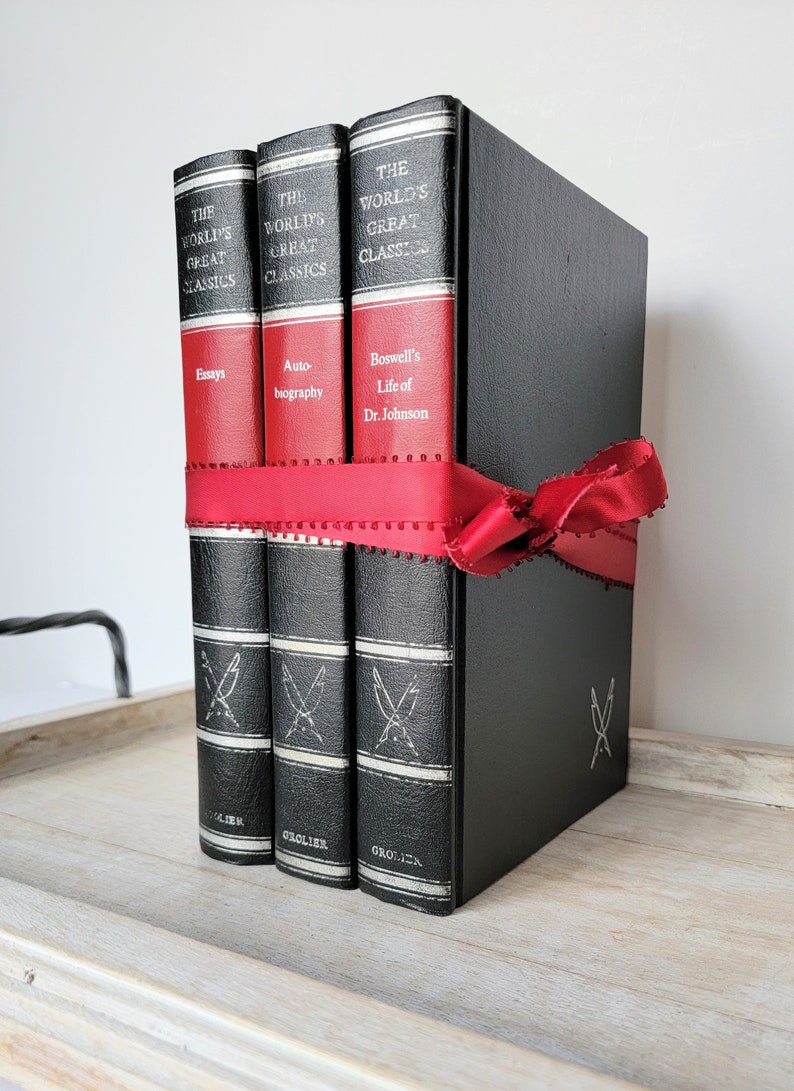 May include: Three black leather-bound books with red spines and gold lettering. The books are titled "The World's Great Classics" and feature the titles "Essays", "Autobiography", and "Boswell's Life of Dr. Johnson". The books are tied together with a red ribbon.