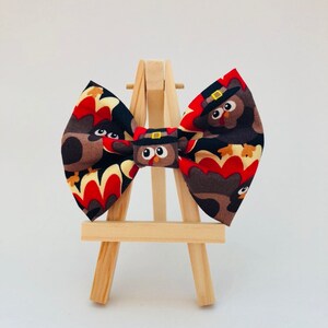 May include: A fabric bow tie featuring a repeating pattern of cartoon turkeys wearing black pilgrim hats with yellow buckles. The turkeys are brown with red and yellow tail feathers against a black background. The bow tie is centered on a wooden stand.