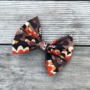 May include: A fabric bow tie featuring a repeating pattern of cartoon turkeys wearing black pilgrim hats. The design includes brown, orange, red, and yellow colors set against a black background, with the turkeys displaying large white eyes.