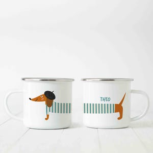 Op de afbeelding: Twee witte emaille mokken met een teckel ontwerp. Eén mok heeft een teckel met een baret en een gestreept shirt. De andere mok heeft de naam "THEO" en een teckelstaart die uit het gestreepte shirt steekt.