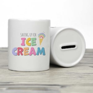 Può includere: Salvadanaio in ceramica bianca con uno slot in cima. La parte anteriore del salvadanaio ha il testo "Saving up for Ice Cream" in lettere colorate con una grafica di cono gelato arcobaleno.