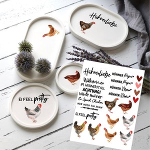Könnte beinhalten: Ein Set aus vier weißen Keramiktabletts mit Hühnerdesign. Die Tabletts sind oval geformt und haben einen erhöhten Rand. Die Tabletts sind mit Aquarell-Illustrationen von Hühnern und dem Text "Hübnerliebe" und "Ei Feel Pretty" verziert.