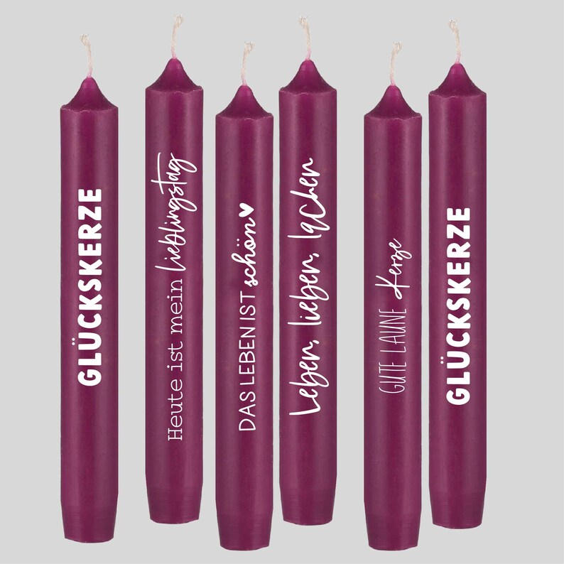 May include: Set of six purple candles with white text. The text on the candles reads: "GLUCKSKERZE", "Heute ist mein Lieblingstag", "DAS LEBEN IST sch&ouml;n", "Leben, lieben, lachen", "GUTE LAUNE Kerze", and "GL&Uuml;CKSKERZE".