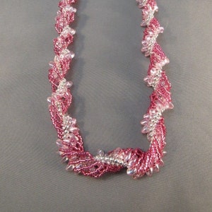 May include: A handmade necklace featuring a spiral design of pink and silver beads. The necklace has a silver clasp and is displayed against a gray background. The beads are arranged in a twisted pattern, creating a textured look.
