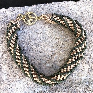 Puede incluir: Un brazalete trenzado con cuentas verdes y doradas alternas. El brazalete tiene un cierre dorado.