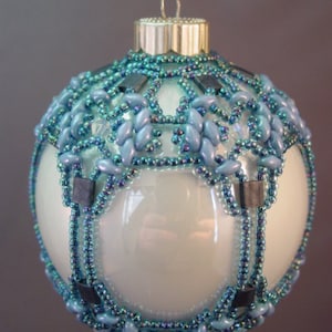 Puede incluir: Un adorno de Navidad de vidrio blanco con un diseño de cuentas azules y negras. El adorno es redondo y tiene una tapa plateada en la parte superior.