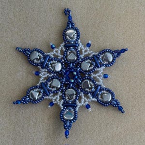 K&ouml;nnte beinhalten: Ein sechsstrahliger Sternschmuck aus komplexer Perlenarbeit. Das Design kombiniert kleine wei&szlig;e Saatperlen, dunkelblaue zylindrische Perlen und gr&ouml;&szlig;ere, glatte, metallisch silberfarbene herzf&ouml;rmige Perlen, die in einem symmetrischen Muster angeordnet sind.