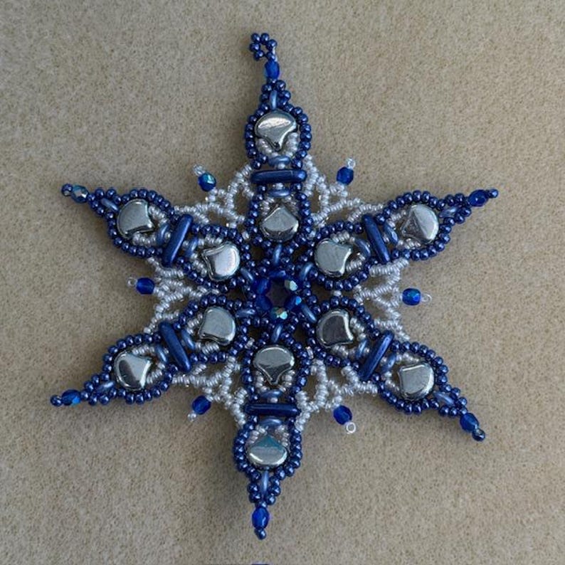 K&ouml;nnte beinhalten: Ein sechsstrahliger Sternschmuck aus komplexer Perlenarbeit. Das Design kombiniert kleine wei&szlig;e Saatperlen, dunkelblaue zylindrische Perlen und gr&ouml;&szlig;ere, glatte, metallisch silberfarbene herzf&ouml;rmige Perlen, die in einem symmetrischen Muster angeordnet sind.