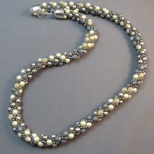May include: A silver and cream beaded necklace with a twisted design. The necklace is made of small, round beads in a variety of shades of silver and cream.