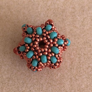 May include: A star-shaped bead crafted from metallic copper-colored seed beads and interspersed with smooth, opaque turquoise-colored beads. The design features a central circular opening and a textured, multi-layered surface construction.