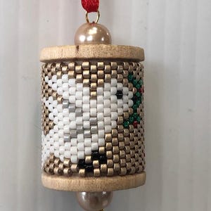 May include: A handmade beaded ornament with a cylindrical shape. The ornament features a design with white, gold, black, and green beads. It has wooden end caps and a red ribbon for hanging. A pearl bead is attached to the top and bottom.