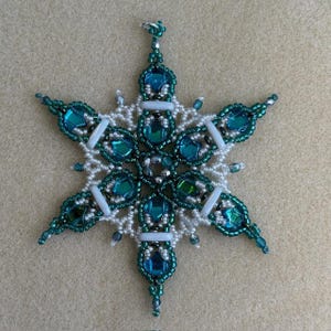 May include: A six-pointed star ornament crafted from intricate beadwork. The design features deep teal and blue faceted glass stones surrounded by small white, dark green, and metallic silver seed beads, with white cylindrical accent beads.