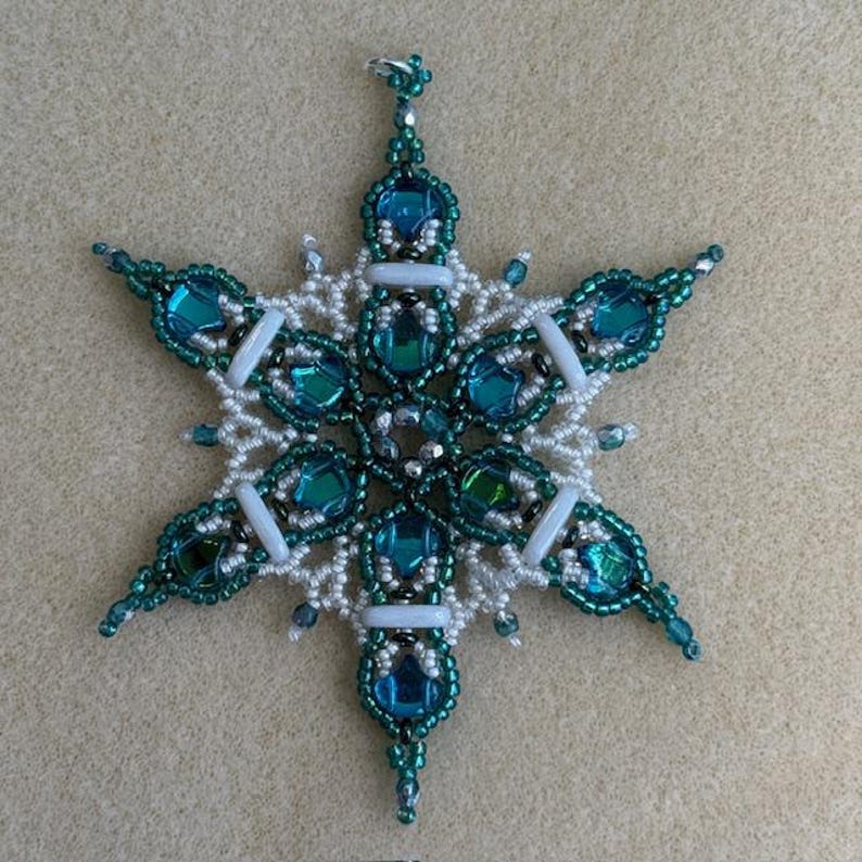 K&ouml;nnte beinhalten: Ein sechsstrahliger Sternschmuck aus komplexer Perlenarbeit. Das Design besteht aus tief t&uuml;rkisfarbenen und blauen facettierten Glassteinen, umgeben von kleinen wei&szlig;en, dunkelgr&uuml;nen und metallisch silbernen Saatperlen sowie wei&szlig;en Zylinderperlen.