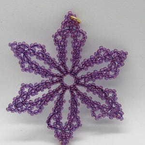 May include: A handcrafted, purple beaded snowflake ornament. The ornament is made of small, round beads strung together to form a six-pointed star shape. A small gold-colored loop is at the top for hanging. The snowflake is a decorative item.