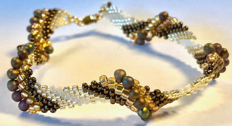 Op de afbeelding: Een kralenarmband met een spiraalvormig ontwerp. De armband is gemaakt met bruine, witte en gouden kralen.