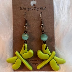 May include: A pair of handmade earrings featuring lime green dragonfly pendants. Each earring has a light blue bead and is attached to a brown earring hook. The earrings are displayed on a brown card with the text "Designs By Red" and "Nature Inspired Designs".