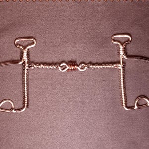 May include: A silver-toned metal horse bit with a twisted wire center and curved side pieces. The bit has a symmetrical design with a loop at the top of each side and a series of small loops on the lower portion of each side.