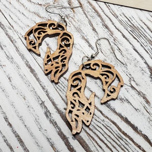 May include: A pair of wooden earrings with a delicate, intricate design. The earrings are shaped like a stylized wave or a flowing pattern. The earrings are made of light brown wood and have silver hooks.
