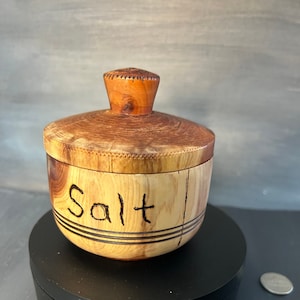 Peut inclure: Un salière en bois avec un couvercle et un petit bouton. Le mot "Salt" est gravé sur le côté du récipient. La salière est faite de bois brun clair et foncé.