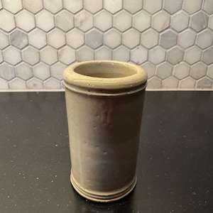 May include: A cylindrical, stoneware jar with a light brown glaze. The jar has a rounded rim and base, and a smooth, unadorned body. The jar is likely vintage or antique, and would be suitable for kitchen or home decor.
