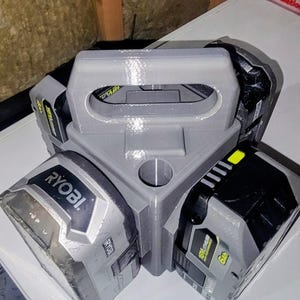 May include: A gray 3D-printed plastic battery holder with a central handle, designed to hold three Ryobi 18V lithium-ion batteries. The holder features a triangular structure with slots for the batteries, which are gray and black with yellow accents and the Ryobi brand name printed on the front.