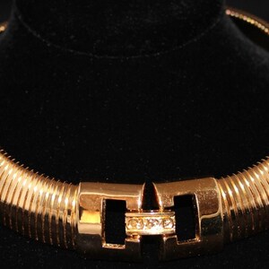 Op de afbeelding: Een gouden chokerketting met een textuur, geribbeld ontwerp. De ketting heeft een eenvoudige, rechthoekige sluiting met heldere strass stenen.