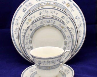 Può includere: Un servizio di stoviglie formali con una tazza e un piattino, insieme a diversi piatti di varie dimensioni. I pezzi in porcellana bianca sono adornati da un delicato motivo floreale blu e dettagli dorati, creando un'estetica elegante.