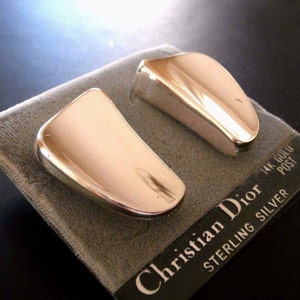 Puede incluir: Un par de pendientes dorados pulidos con un diseño rectangular curvo. Los pendientes se muestran sobre una tela gris con una etiqueta que dice "Christian Dior STERLING SILVER 14K GOLD POST."